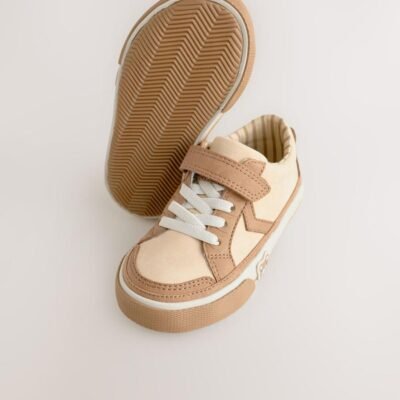 These adorable low-top trainers blend soft textures and neutral tones, featuring a large star graphic and multiple charming bear motifs for a fun, stylish look, secured by a single hook-and-loop strap. Brand: Next UK Mixed Media Upper: The low-cut body combines smooth light beige/cream synthetic material on the side and vamp, with tan/brown faux suede/nubuck overlays on the toe cap, eyelet area, and strap. Star Graphic Detail: The side panel features a large star-shaped graphic in the smooth light beige material, sharply defined by the darker tan stitching and overlay edges. Hybrid Closure: The shoe includes fixed, wide cream/white laces for a traditional look, secured by a single, adjustable hook-and-loop (Velcro) strap made of the tan faux suede. Multiple Bear Motifs: The heel patch features a sweet embroidered bear face. A second, small embossed bear face graphic is visible on the outer sidewall of the gum rubber sole, adding character. Gum Rubber Sole: The trainers are set on a flat, thick gum/natural colored rubber sole with a classic chevron/zig-zag tread pattern. The sidewall features a subtle contrast stripe. Color: Upper is predominantly Light Beige / Cream with Tan / Brown overlays. Sole is Gum / Natural Brown. Lining is Beige and White Striped. A comfortable and charming low-top trainer featuring a multi-tonal neutral upper, a star graphic, and playful bear character accents.