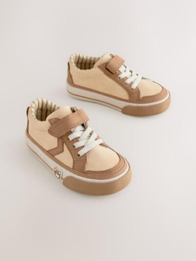 These adorable low-top trainers blend soft textures and neutral tones, featuring a large star graphic and multiple charming bear motifs for a fun, stylish look, secured by a single hook-and-loop strap. Brand: Next UK Mixed Media Upper: The low-cut body combines smooth light beige/cream synthetic material on the side and vamp, with tan/brown faux suede/nubuck overlays on the toe cap, eyelet area, and strap. Star Graphic Detail: The side panel features a large star-shaped graphic in the smooth light beige material, sharply defined by the darker tan stitching and overlay edges. Hybrid Closure: The shoe includes fixed, wide cream/white laces for a traditional look, secured by a single, adjustable hook-and-loop (Velcro) strap made of the tan faux suede. Multiple Bear Motifs: The heel patch features a sweet embroidered bear face. A second, small embossed bear face graphic is visible on the outer sidewall of the gum rubber sole, adding character. Gum Rubber Sole: The trainers are set on a flat, thick gum/natural colored rubber sole with a classic chevron/zig-zag tread pattern. The sidewall features a subtle contrast stripe. Color: Upper is predominantly Light Beige / Cream with Tan / Brown overlays. Sole is Gum / Natural Brown. Lining is Beige and White Striped. A comfortable and charming low-top trainer featuring a multi-tonal neutral upper, a star graphic, and playful bear character accents.